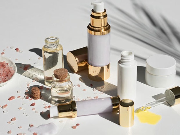 Botecitos blancos y dorados con productos para el cuidado facial sobre un fondo blanco. También hay granos de sal rosa.