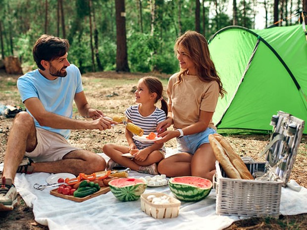Una familia de un padre, una madre y una niña haciendo un picnic en el campo. De fondo se ve una tienda de campaña verde.