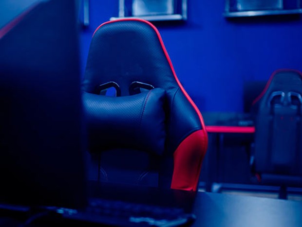 Silla de gaming en primer plano tras un monitor de PC. La silla es negra y roja y tiene un cojín. En el fondo se ve otra silla igual.