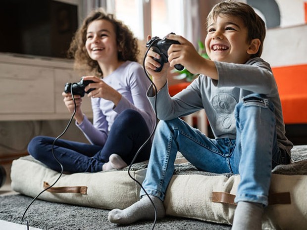 Un niño y una niña sonrientes jugando a videojuegos mientras están sentados en un cojín en el suelo de un salón