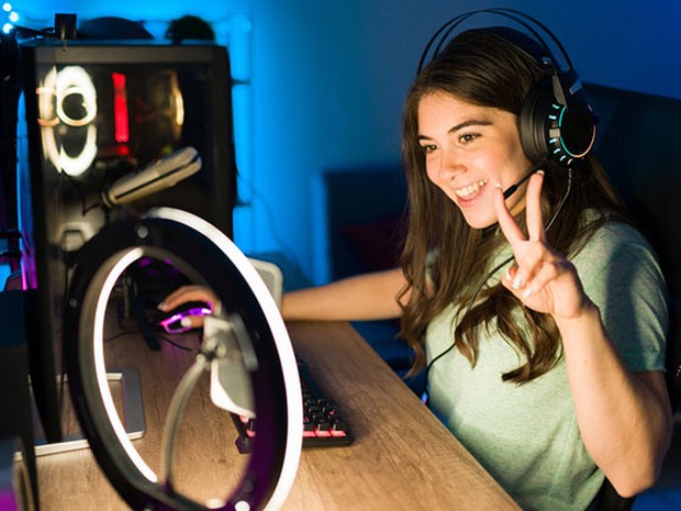 Chica joven mirando a un smartphone iluminada con un aro de luz. La chica lleva unos cascos de gamer y está haciendo el símbolo de la victoria con la mano izquierda.