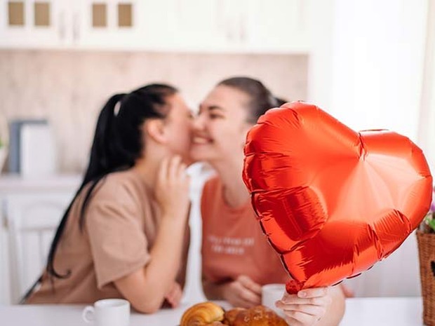 Una pareja joven de dos mujeres dándose un beso en la mejilla mientras desayunan en su cocina. Una de ellas sujeta en primer plano un globo en forma de corazón.