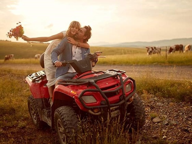Pareja joven de un chico y una chica. El chico está conduciendo un quad rojo y dándole un beso en la mejilla a la chica que le sujeta desde atrás.