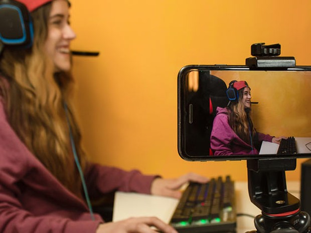 Smartphone sujetado en un trípode y grabando a una chica haciendo streaming. La chica lleva unos cascos de gamer y una sudadera magenta.