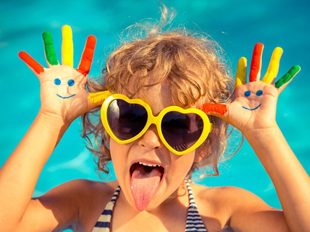Una niña con el pelo corto y rizado en una piscina con unas gafas de sol amarillas en forma de corazón y las manos pintadas de colores. Tiene las manos a la altura de la cabeza y saca la lengua.