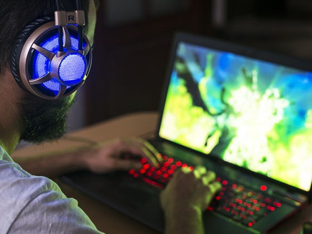 portátil de gaming con las teclas iluminadas en rojo y un vídeojuego en pantalla. Usándolo hay un hombre joven con barba y unos cascos iluminados en azul