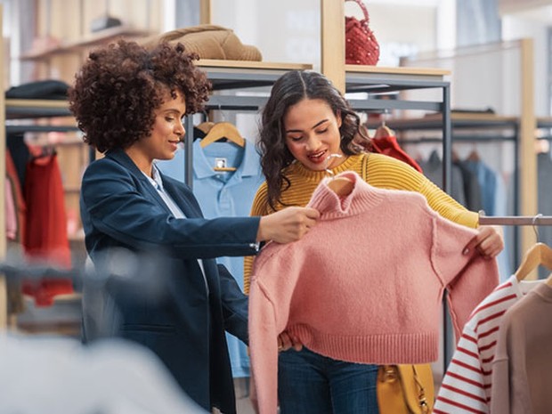Mujer adulta con el pelo rizado y traje de chaqueta enseñándole un jersey rosa a una mujer joven en una tienda de ropa