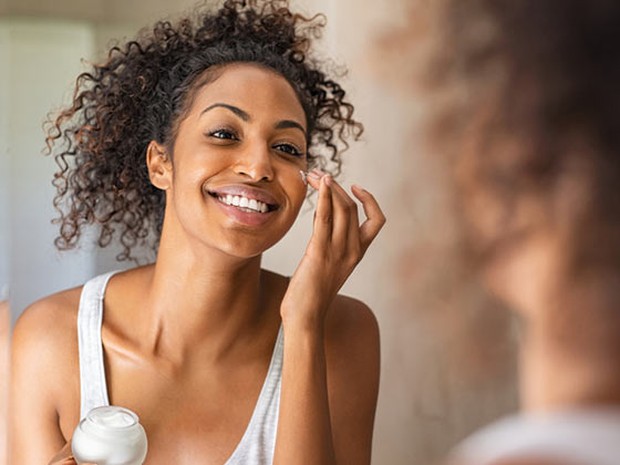 Mujer mirándose a un espejo con un bote de crema facial y echándosela en la cara