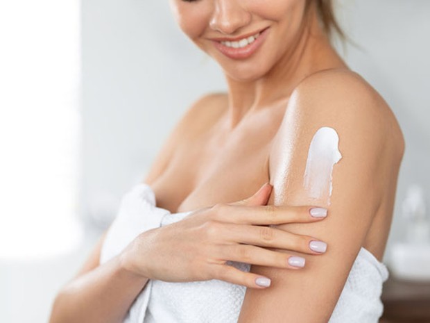Mujer joven echándose crema aftersun en el brazo después de salir de la ducha envuelta en una toalla blanca