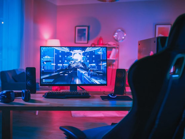 Set de gamer con luces azules y rosas. Silla de gamer en primer plano, monitor con un videojuego puesto encima de una mesa de madera en la que también hay unos cascos y unos altavoces