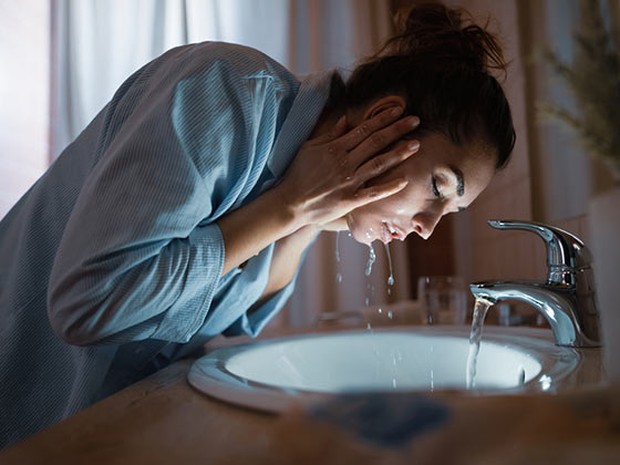 Mujer lavándose la cara en el lavabo de su casa. Lleva un pijama azul y el pelo recogido en un moño.