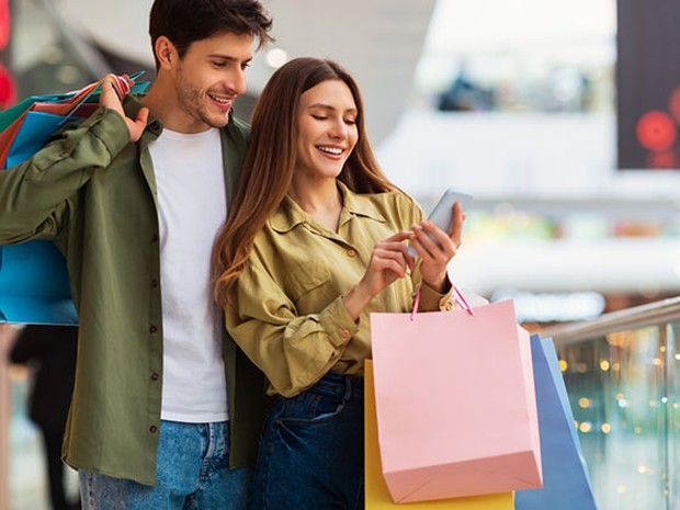Pareja joven en un centro comercial mirando el smartphone de ella y con bolsas de cartón de colores en las manos