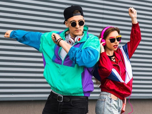 Chica joven y chico joven bailando delante de una verja plateada. Van vestidos con sudaderas retro. La de él es verde, morada y azul y la de ella es granate, azul y blanca.