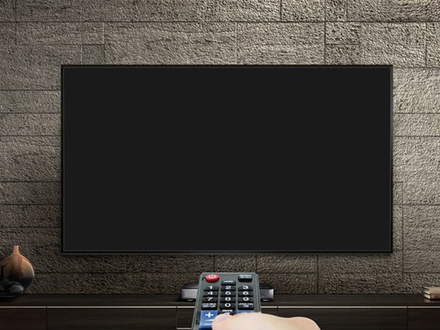 Televisión plana de pared sobre una pared de ladrillo gris. Mando a distancia en primer plano con una mano pulsándolo.