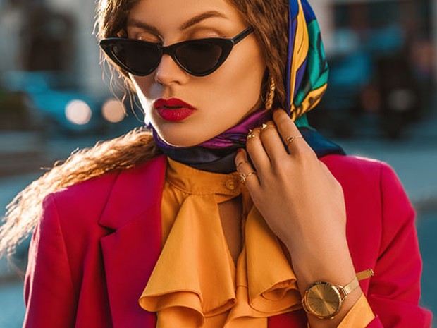 Mujer joven con un pañuelo retro en la cabeza, unas gafas de sol con forma triangular, labios pintados de rojo a juego de su blazer, camisa con cuello con volantes naranjas y accesorios dorados (reloj y anillos)