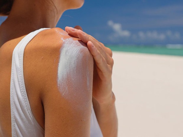 Mujer echándose crema solar en el hombro. Lleva un bañador blanco y de fondo se ve una playa paradisiaca.
