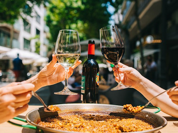 Dos personas brindando con una copa de vino tinto y una de blanco y comiendo una paella con cuchara