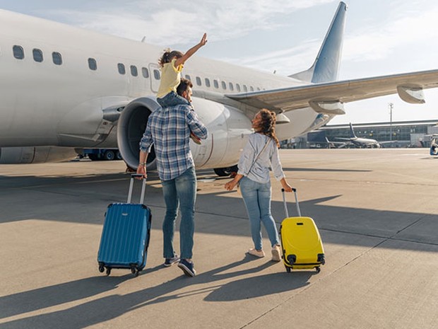 Familia de padre, madre e hija caminando hacia un avión. La madre lleva una maleta amarilla y el padre una azul. La hija va en los hombros de su padre.