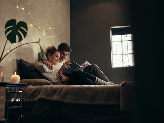 Pareja joven de chico y chica tumbados en una cama leyendo juntos un libro. La habitación está iluminada por una vela y una guirnalda de lucecitas.
