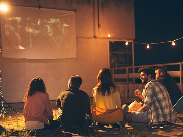 Grupo de 5 amigos sentados en el suelo de una terraza viendo algo en un proyector. La terraza está iluminada con lucecitas.