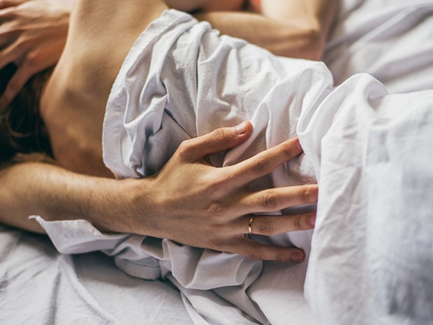Pareja abrazada entre las sábanas. En primer mano se ve una mano masculina con un anillo de oro en el dedo anular. La mano está apoyada en la espalda de una mujer joven.