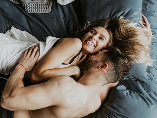 Pareja joven tumbados abrazados en la cama. Ella sonríe mirando hacia el techo y él sonríe mirándola a ella.