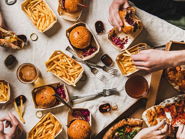 Mesa llena de hamburguesas, patatas fritas y dos pizzas. Hay manos cogiendo la comida.