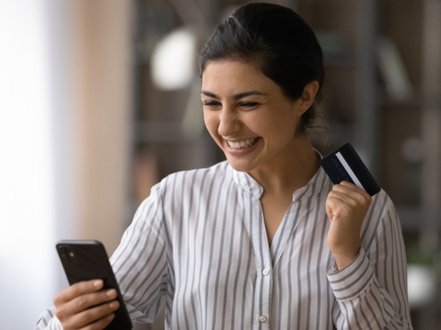 Mujer joven muy sonriente mirando su smartphone y con una tarjeta de crédito en la otra mano