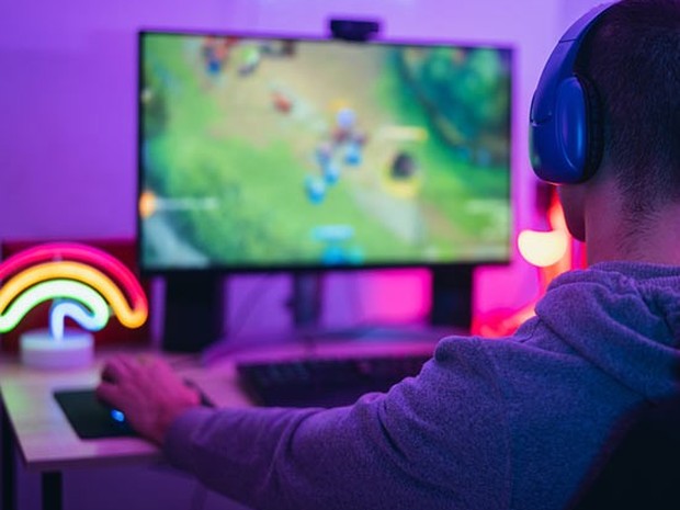 Chico joven jugando al LOL. El chico lleva unos cascos de gamer azul y su habitación está iluminada con luces led moradas, rosas, amarillas y azules