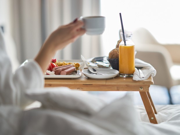 Desayuno de hotel en la cama. En la bandeja de madera se ve un plato con huevos revueltos, tomate y salchichas. También hay una magdalena y un zumo de naranja. Además, la mano de una mujer está cogiendo una taza de café.