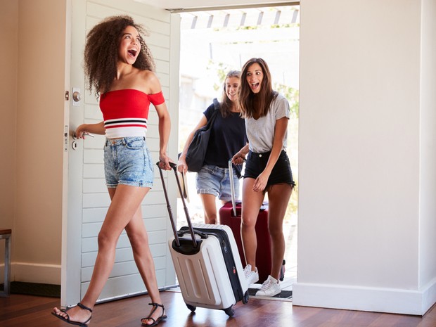 Tres chicas jóvenes entrando en un apartamento vacacional. La primera lleva una maleta de ruedas blanca y la segunda una roja.