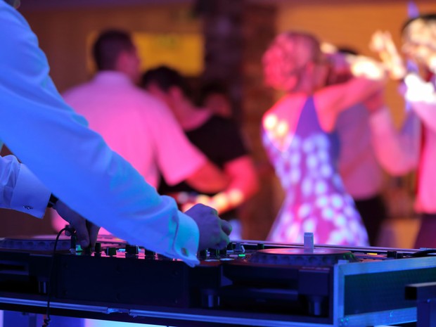 DJ con su mesa de mezclas iluminado por una luz azul. En el fondo se ve a gente bailar iluminados por luces rosas.