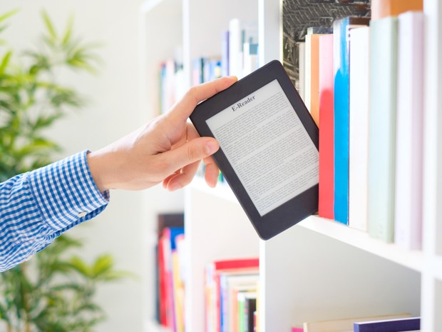 La mano de una persona está cogiendo un eBook de una estantería llena de libros. El eBook está encendido y hay un texto en pantalla.