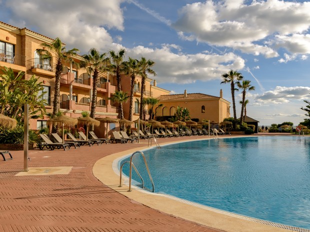 Imagen de la piscina de un hotel isleño. El hotel se ve al fondo con una fachada amarilla y delante hay varias palmeras.