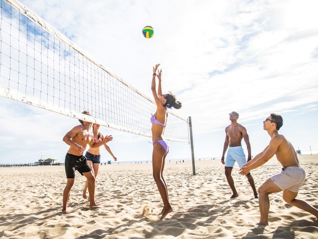 Grupo de dos chicas y tres chicos jóvenes jugando al voley-playa con una pelota verde, amarilla y negra
