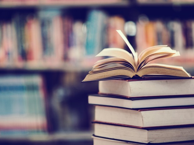 Pila de libros con uno abierto por la mitad encima del todo. De fondo se ven unas estanterías repletas de libros difuminado.