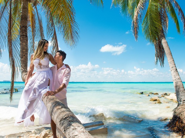 Pareja joven en una playa paradisiaca. Ella lleva un vestido de novia y está sentada encima de una palmera. Él lleva una camisa rosa palo y rodea a ella con su brazo.