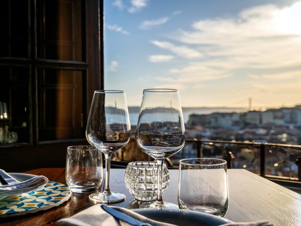 Mesa puesta para cenar con dos copas de vino y dos vasos. De fondo se ve una ciudad costera y el mar a través de una ventana abierta.