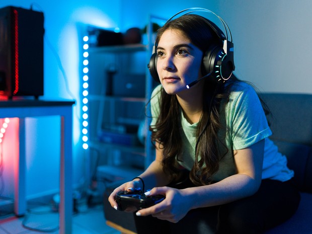 Chica joven con el pelo largo sentada en un sofá jugando a videojuegos. Lleva unos auriculares de gamer con micrófono y de fondo se ven luces led azules y rojas
