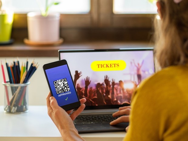 Mujer mirando tickets para un concierto en su portátil. Con una mano sostiene un smartphone donde se ve un código QR con el fondo azul.