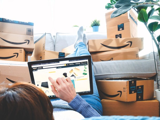 Mujer tumbada en el suelo con los pies en el sofá haciendo un pedido en Amazon. Se ven muchas cajas de Amazon.