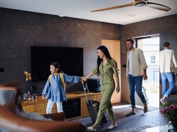 Familia compuesta por padre, madre e hija entrando en lo que parece una habitación de hotel. La niña y la madre llevan una maleta de mano negra y el padre una blanca.