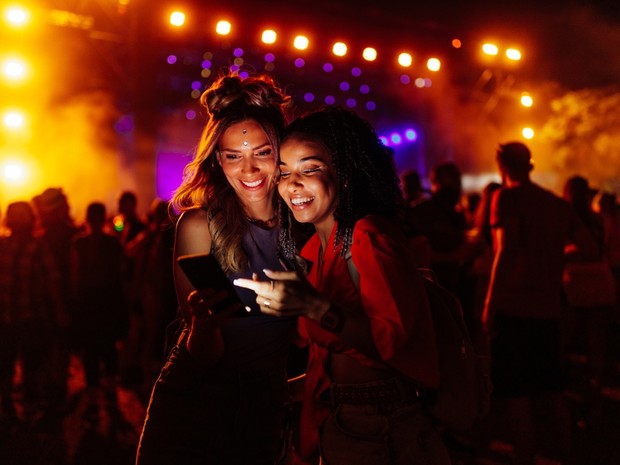Dos chicas jóvenes mirando un smartphone en un festival. De fondo se ven las luces del escenario. Ellas llevan maquillaje festivo.