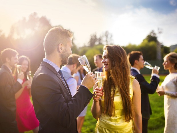 Una pareja en primer plano brindando con champán en una boda. En el fondo se ve a los novios y otros invitados