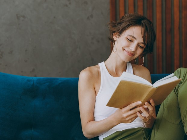 Mujer joven con el pelo corto leyendo un libro con la portada marrón. Está sentada en un sofá de terciopelo azul y sonríe.