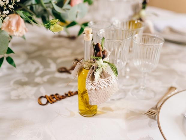 Botellita de aceite con un adorno de tela y plantas. Está encima de una mesa con mantel blanco, copas de cristal y flores