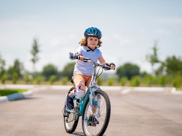 Niño montando en una bicicleta azul en una zona de asfalto en un parque. El niño lleva un casco azul, coderas y rodilleras