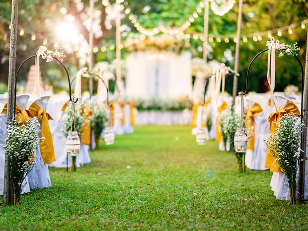 Lugar al aire libre donde se va a celebrar una boda. Se ven guirnaldas de luces, sillas blancas con lazos amarillos, ramos de flores y un altar al fondo.