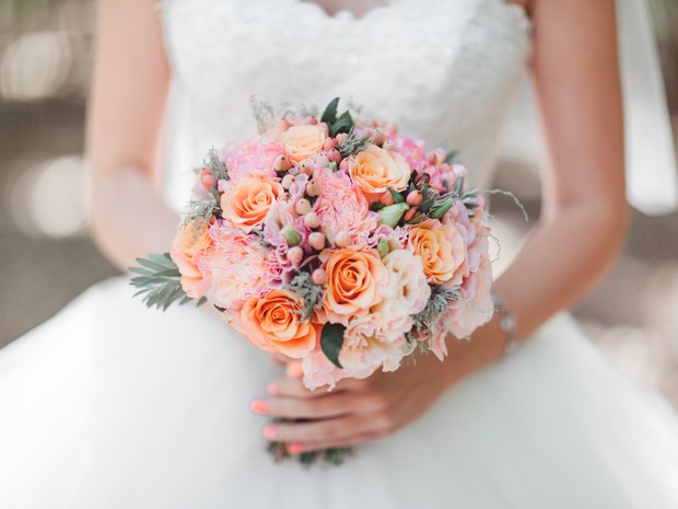 Ramo de flores de novia en tonos rosas y naranjas en primer plano. El ramo lo sujeta la novia, que se ve de fondo su vestido blanco y las uñas pintadas de rosa