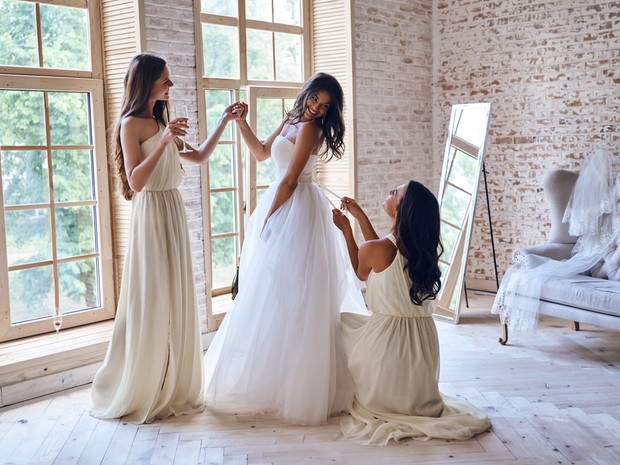 Tres chicas jóvenes. La del centro está vestida de novia y sonríe mientras una de sus amigas le hace un lazo en la cola del vestido blanco. Las dos amigas llevan vestidos crema de damas de honor.